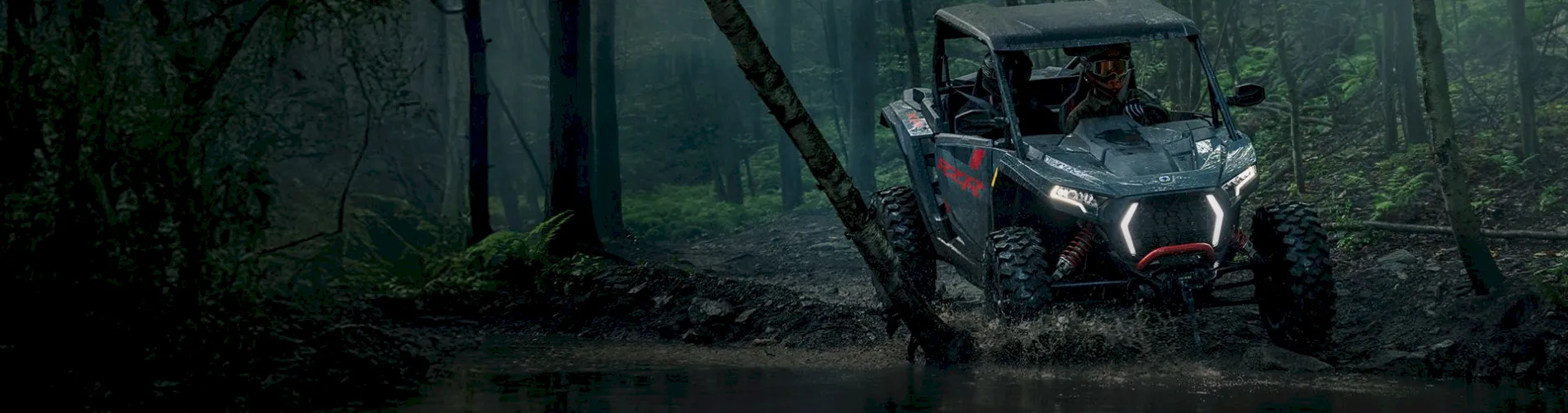 Polaris RZR side-by-side driving through muddy forest trail with rider and illuminated headlights